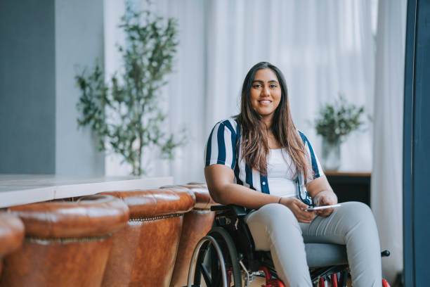 A woman sitting in a wheelchair with a tablet device in her hands. She has long brown hair and is wearing jeans, a white vest top and a blue and white strip short sleeved shirt. There are brown leather tub chairs to her left and a white voile curtain and 2 indoor plants in the background.
